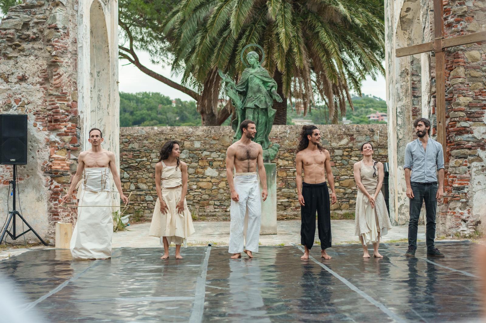 Six performers stand on an outdoor stage after a performance.