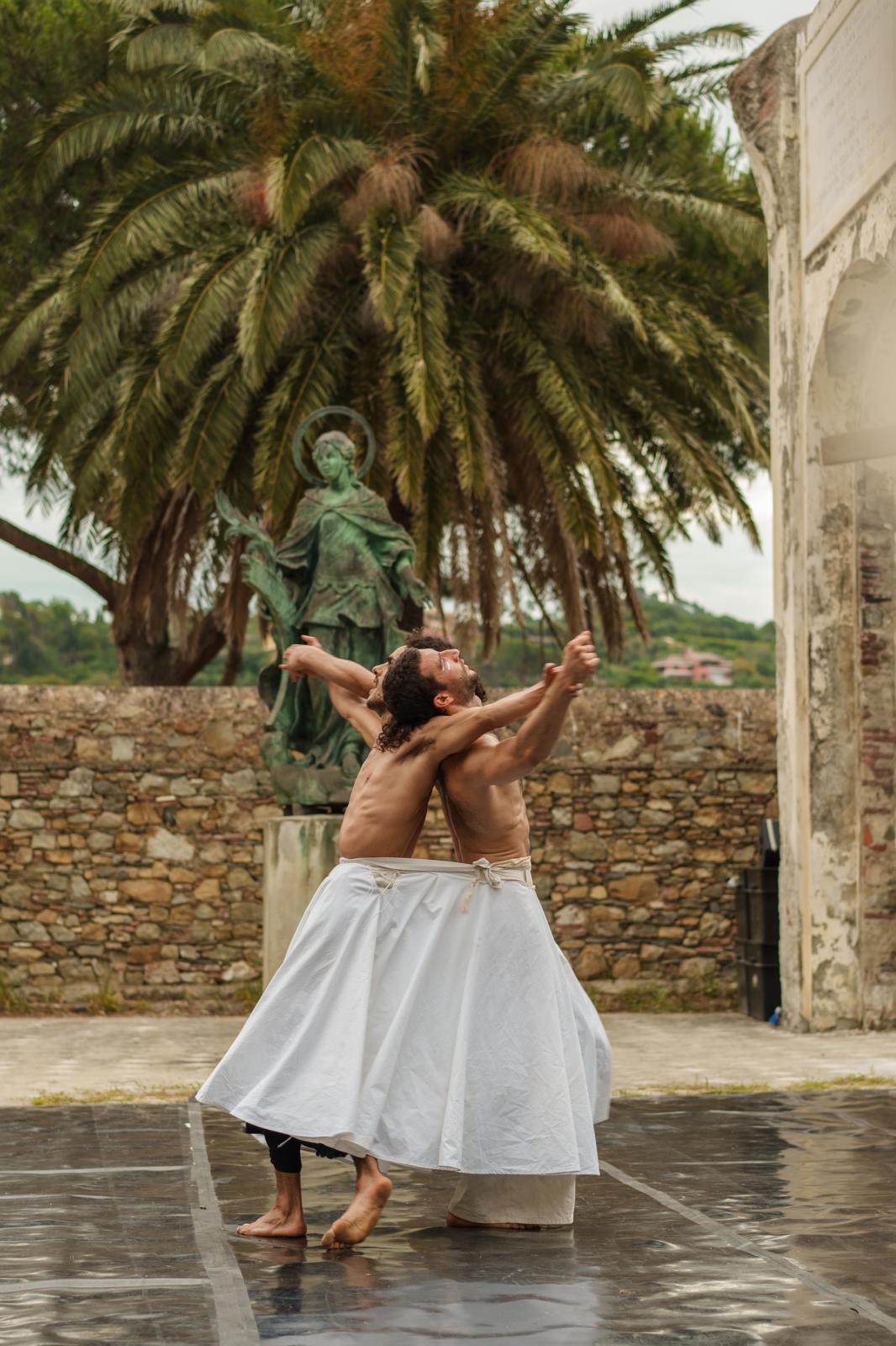 Two dancers in white costumes turn together in front of a palm tree and an old wall.